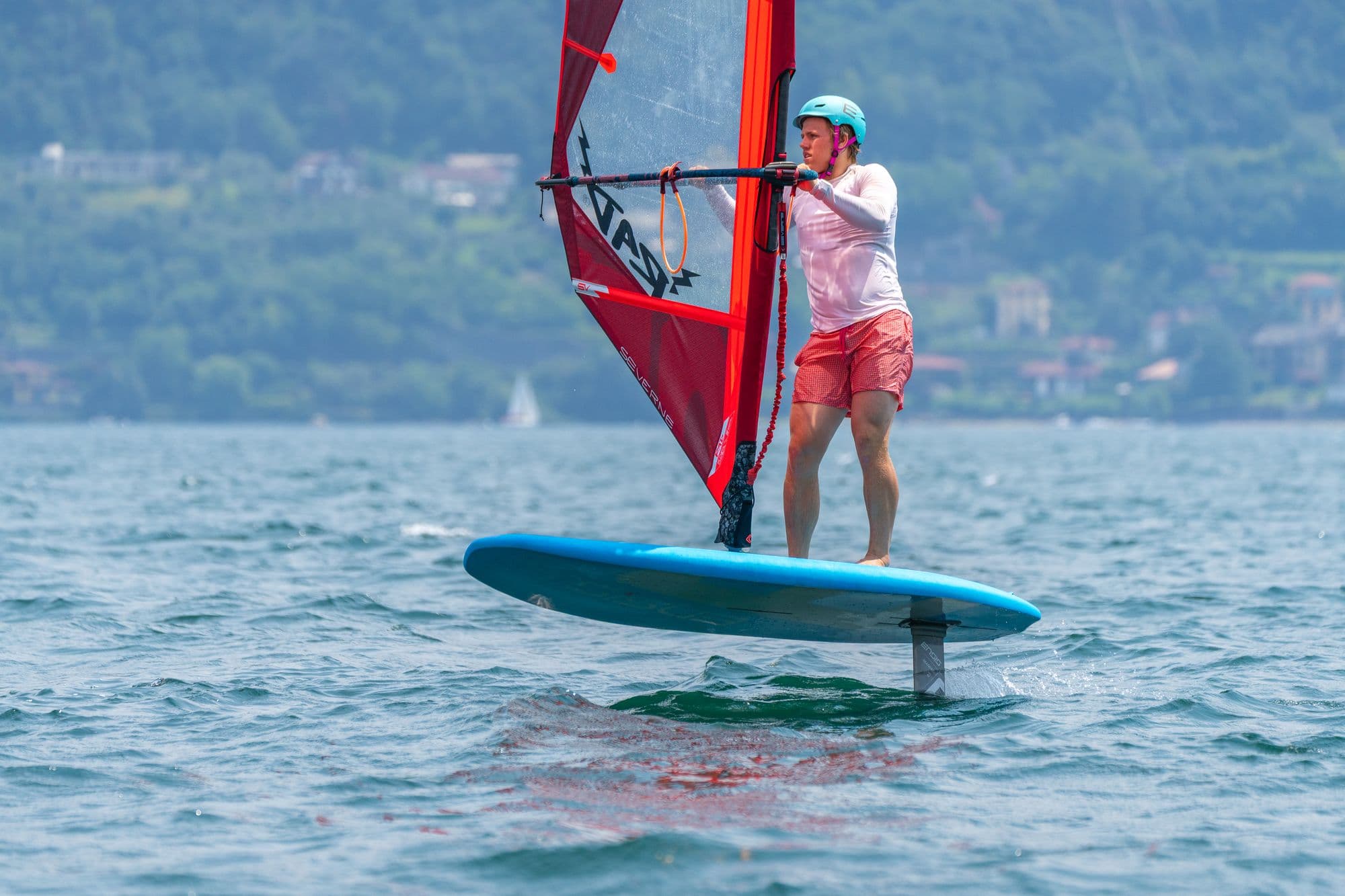 Fahrer aufrecht auf dem Foilboard, rotes Segel, Ufer im Hintergrund