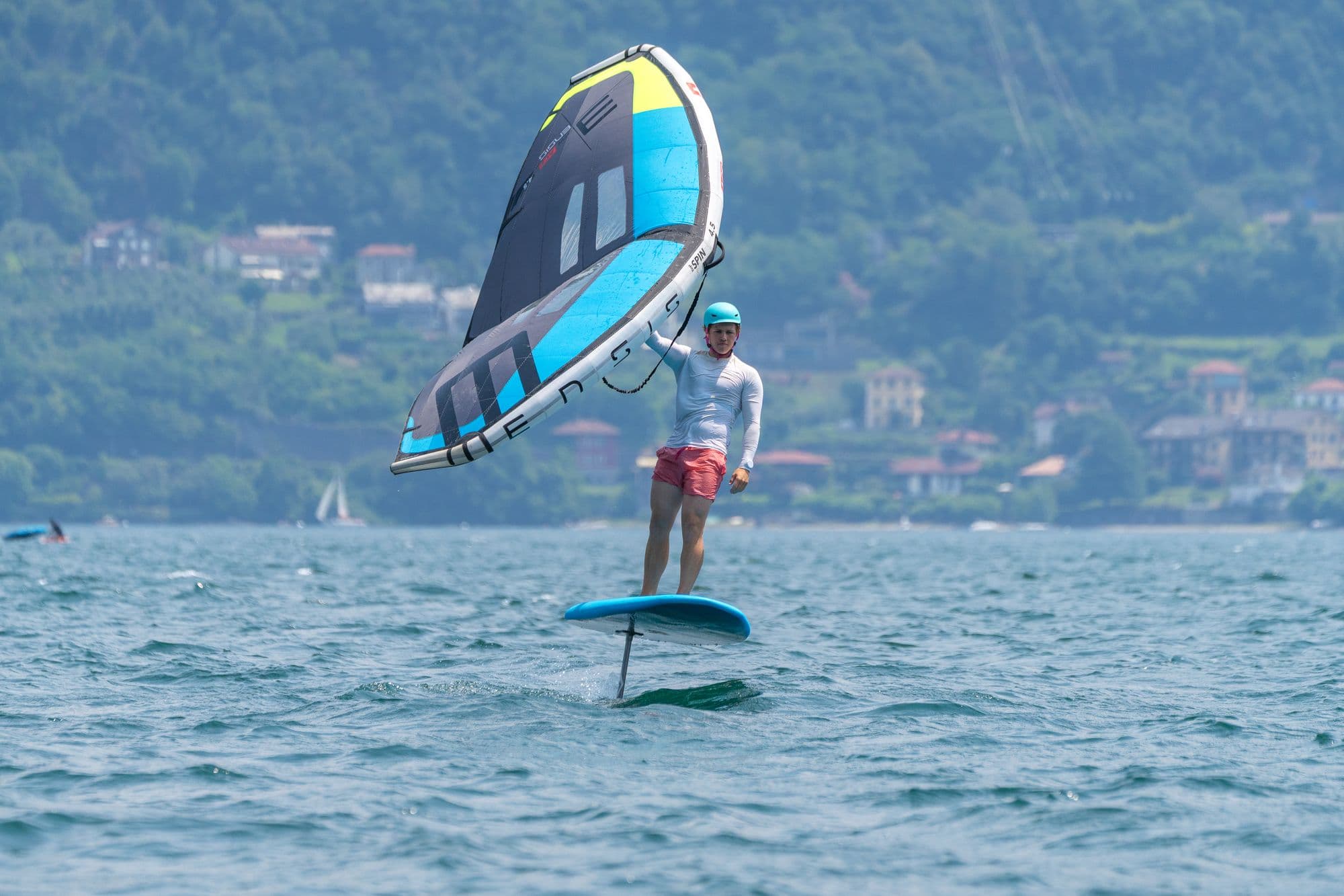 Wingfoiling mit blau-schwarzem ENSIS-Wing, Ufer und Häuser im Hintergrund