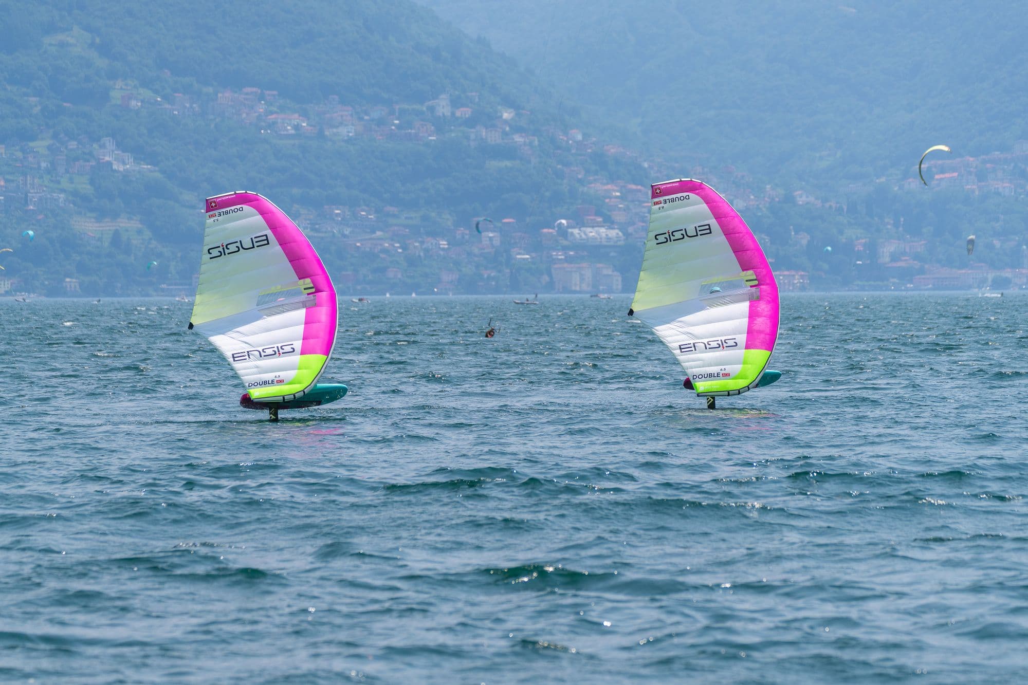 Zwei Wingfoiler in Fahrt, Ufer und Berge im Hintergrund