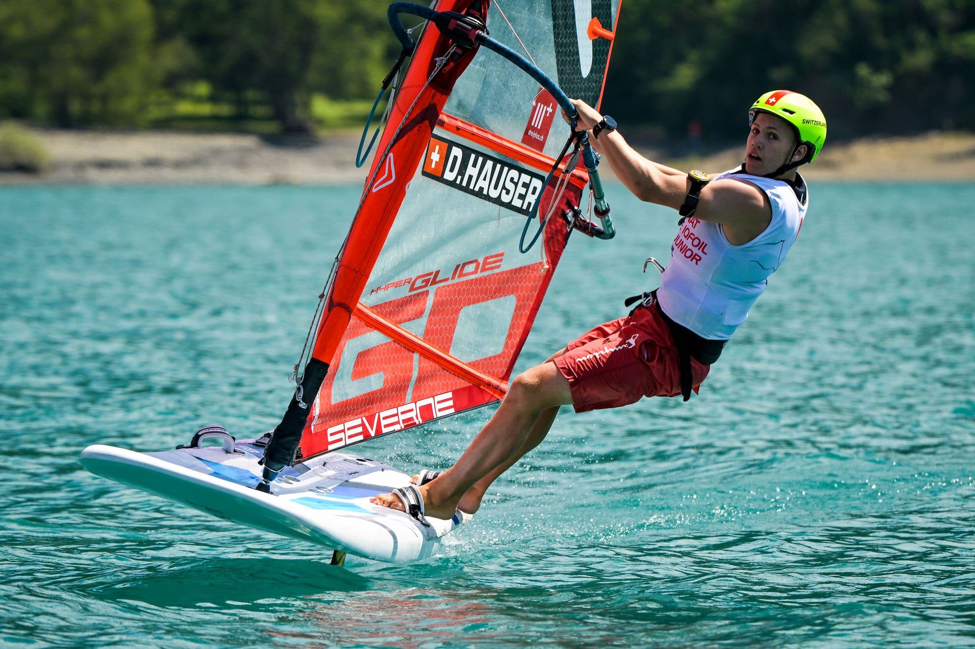 Devin Hauser in roten Boardshorts am Trapez, grünes Ufer im Hintergrund