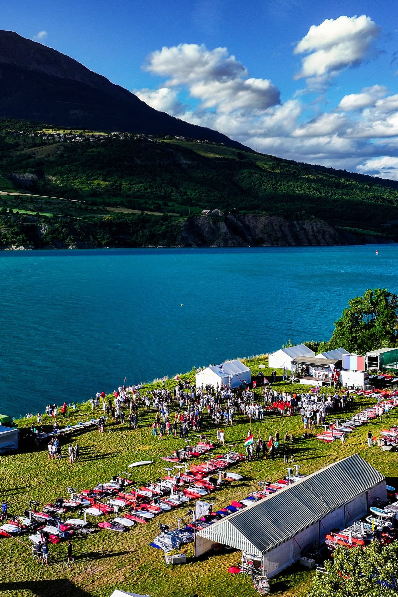 Hochformat über das Regattagelände: Boards in langen Reihen auf der Wiese, dahinter der türkise See und die Berge