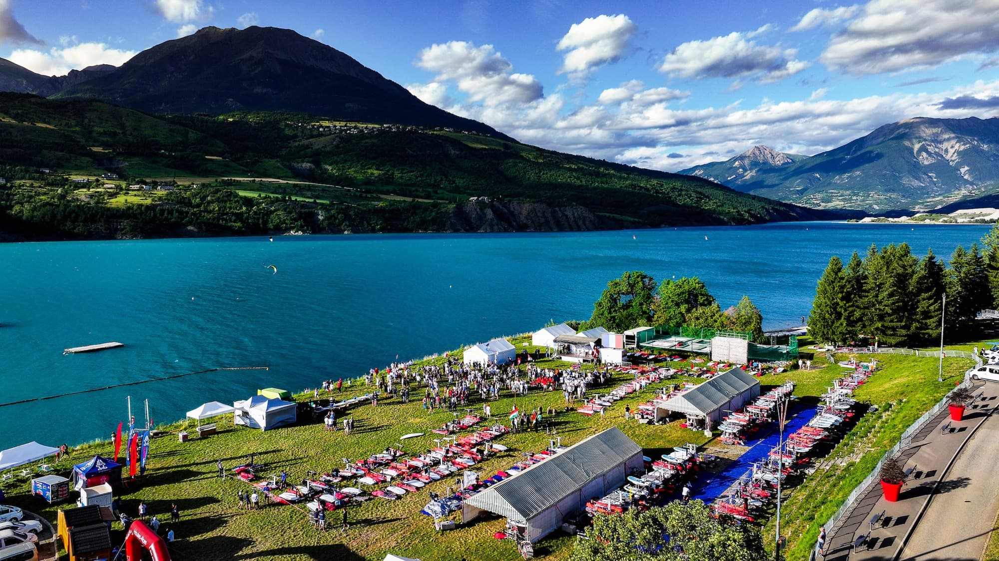 Luftaufnahme des Geländes am See: Zelte, Boards und die versammelten Teams auf der Wiese über dem Wasser
