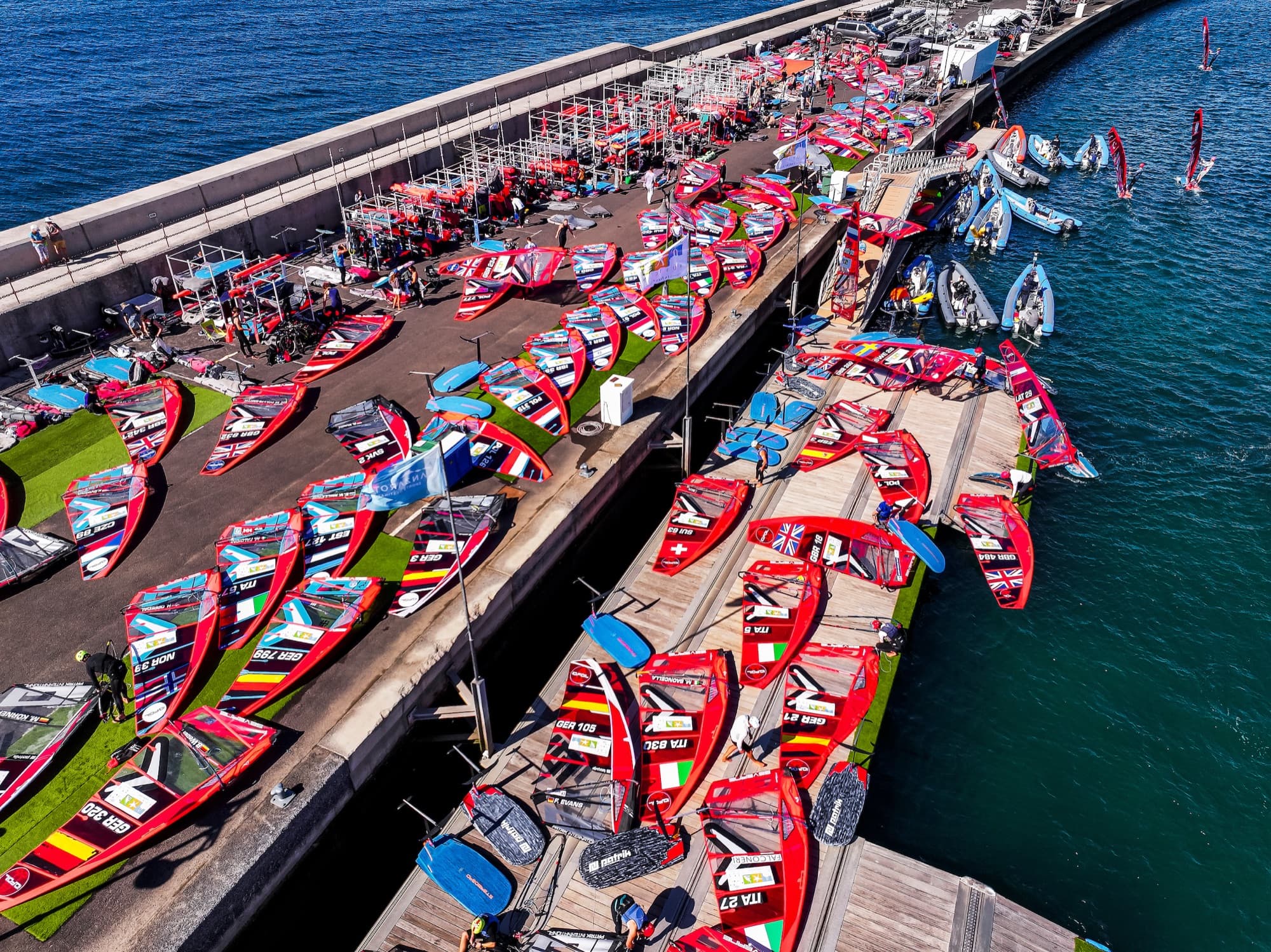 Luftaufnahme des Boat Parks auf der Mole, Segel und Boards reihenweise auf grünen Matten und Stegen