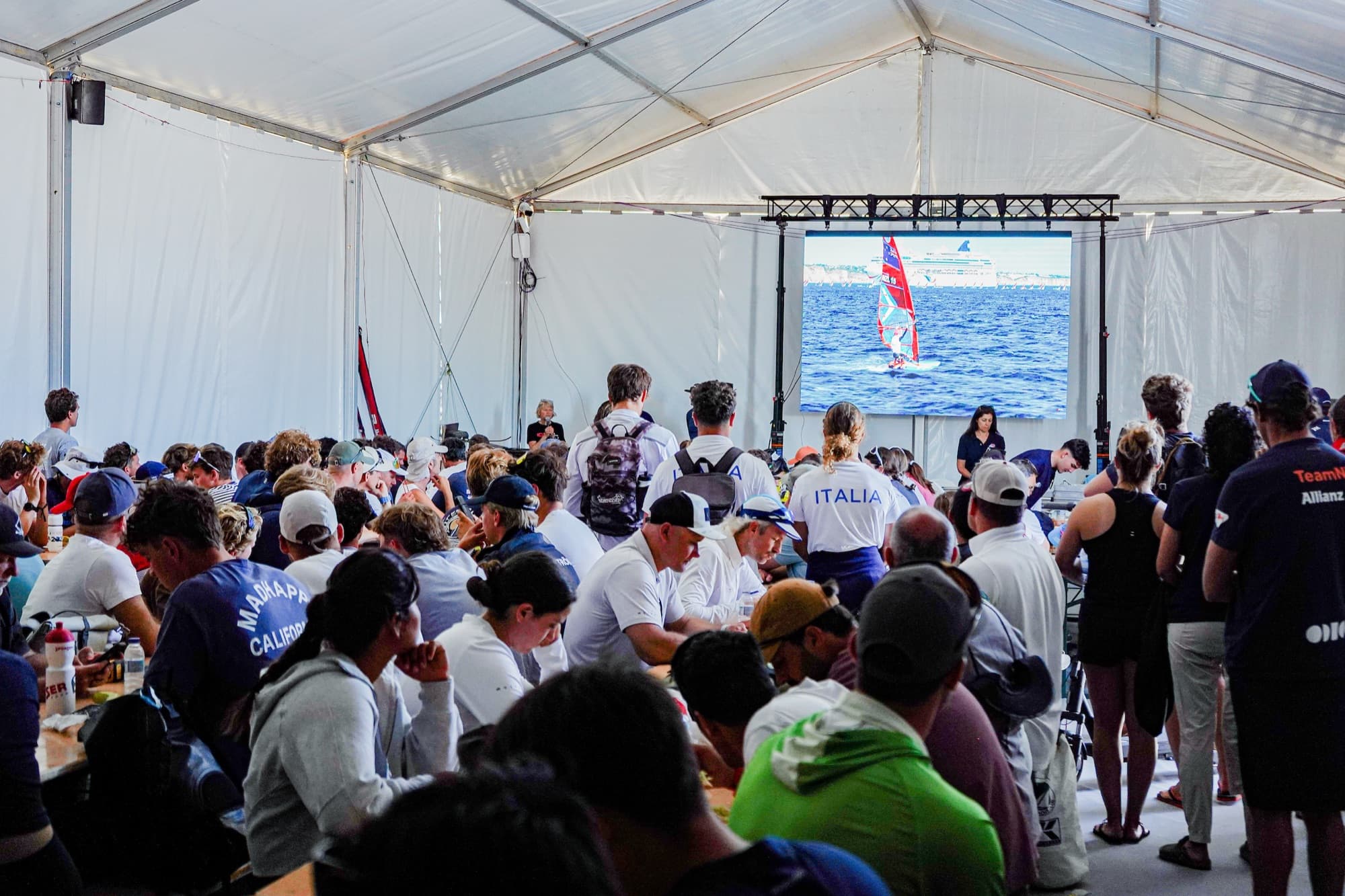 Briefing im Zelt: auf der Leinwand läuft eine Aufnahme vom Wasser, die Teams verfolgen die Ansprache