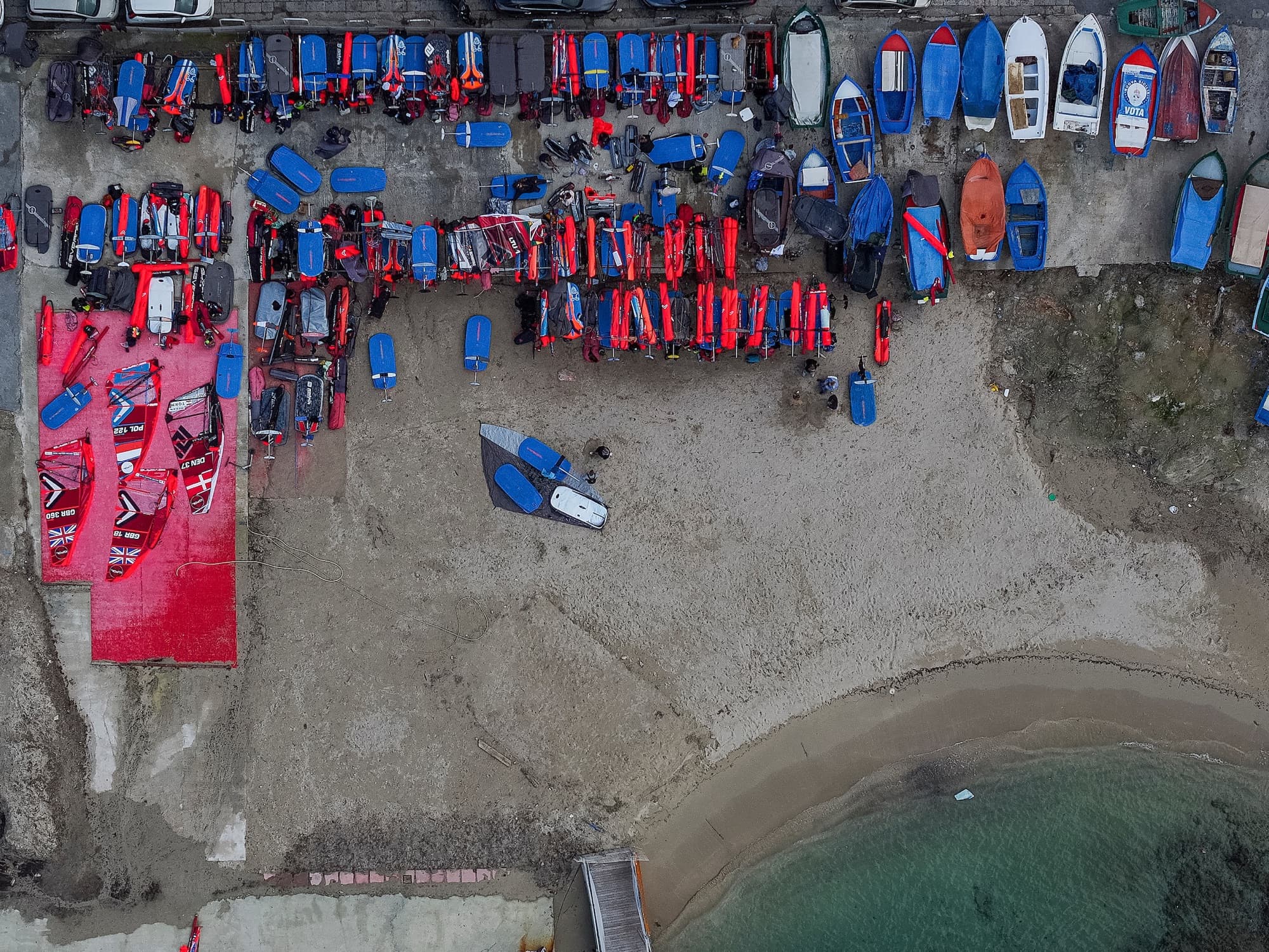 Senkrechte Luftaufnahme über den Materialplatz am Strand, Boards und Segel in langen Reihen neben den Fischerbooten