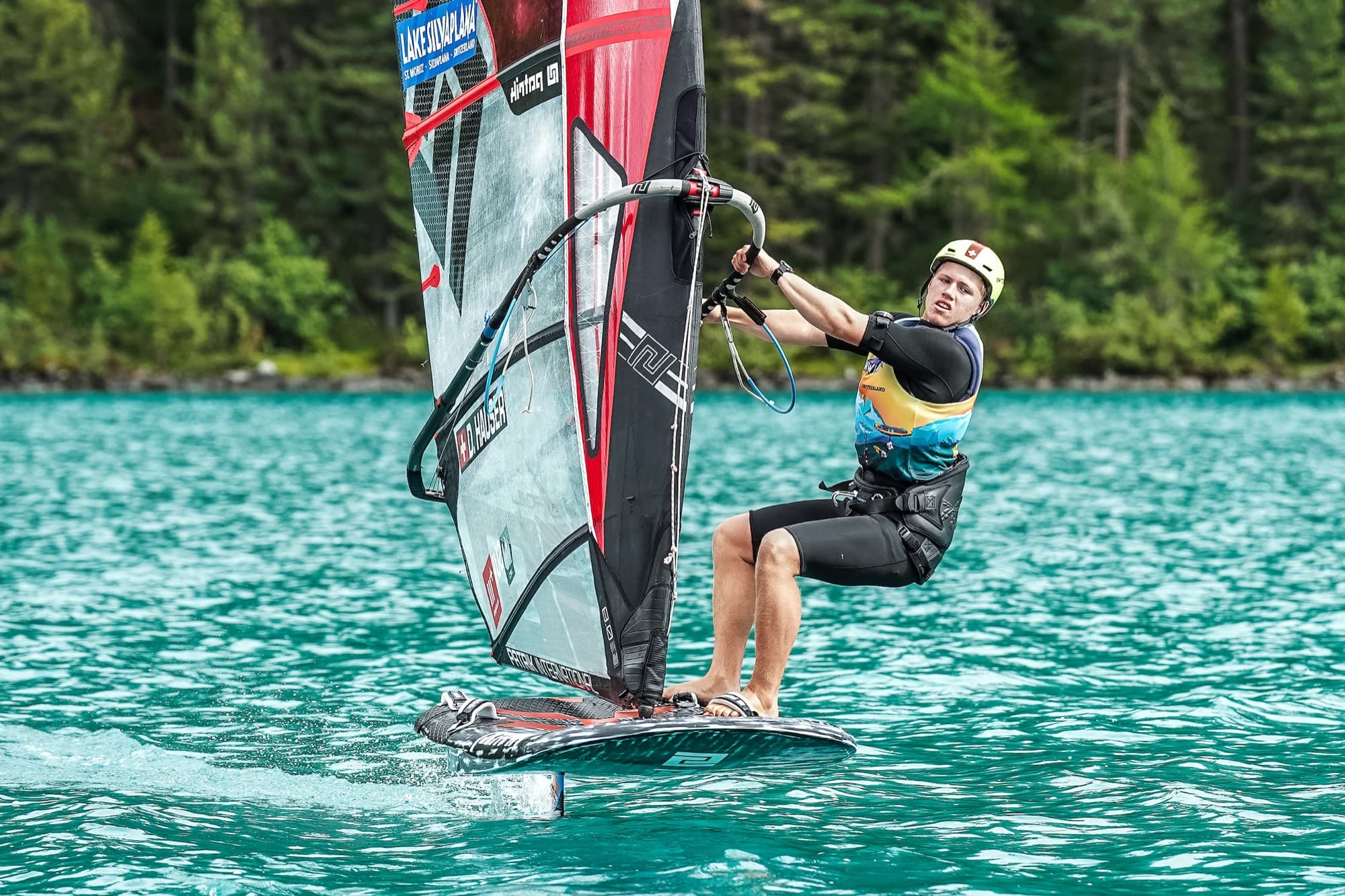Devin Hauser blickt über die Schulter zurück, Namenszug D. HAUSER am Segel, Board auf dem Foil über türkisem Wasser