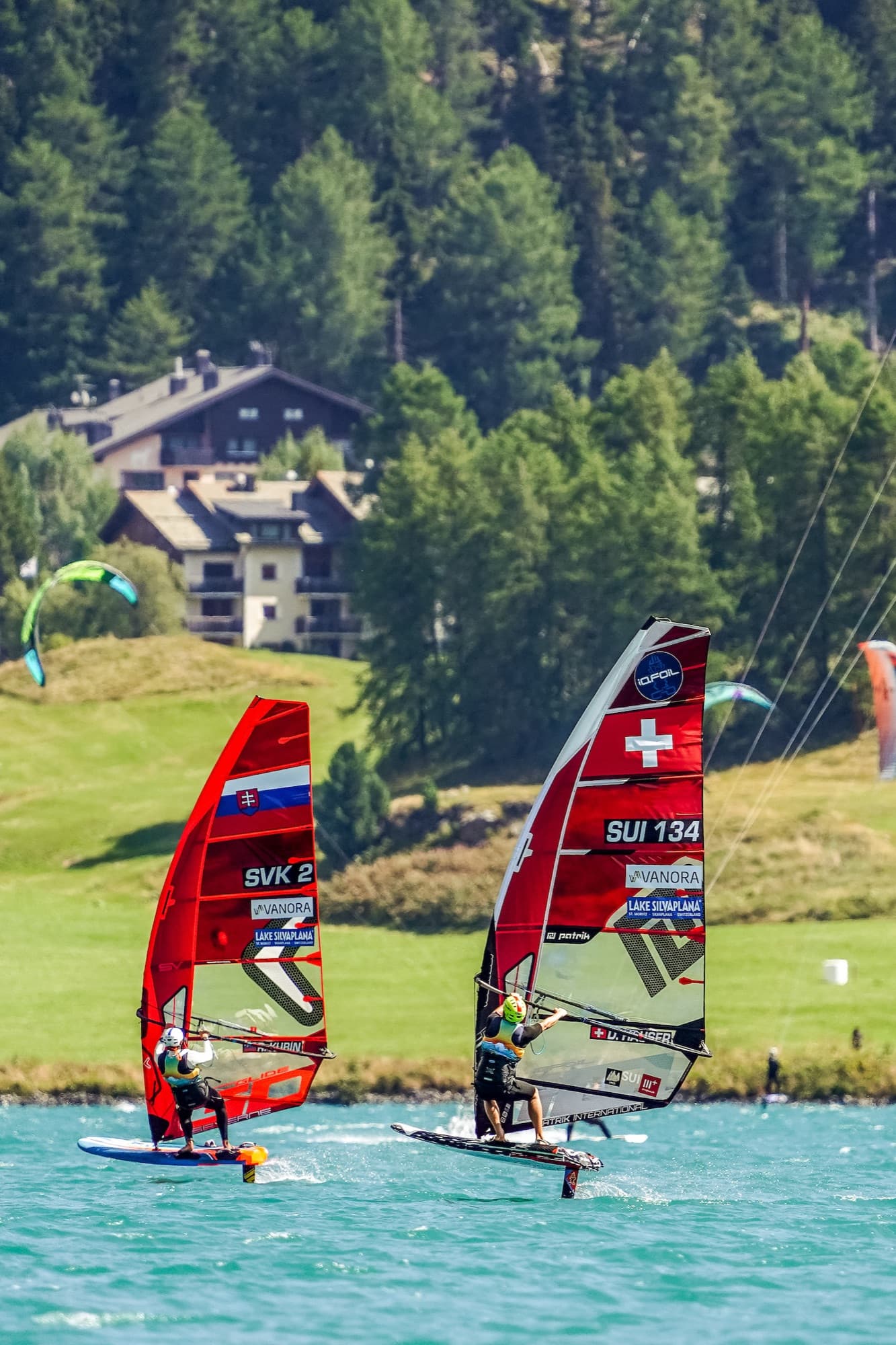 Hochformat: SVK 2 und SUI 134 foilen nebeneinander, am Ufer Chalets und zwei Kites über der Wiese