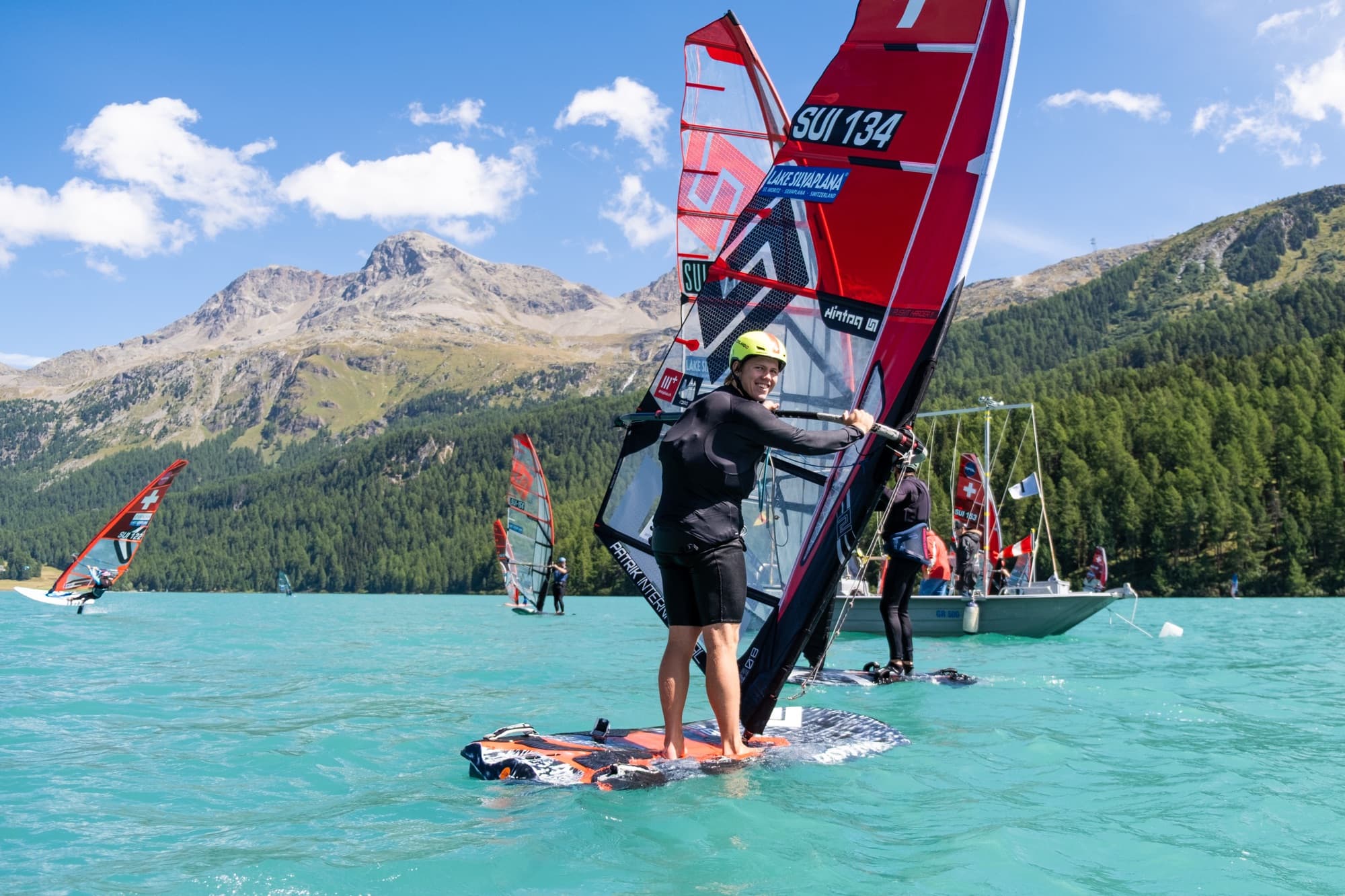 Devin Hauser steht vor dem Start auf dem Board und lacht in die Kamera, Segelnummer SUI 134, Berge und Startboot im Hintergrund