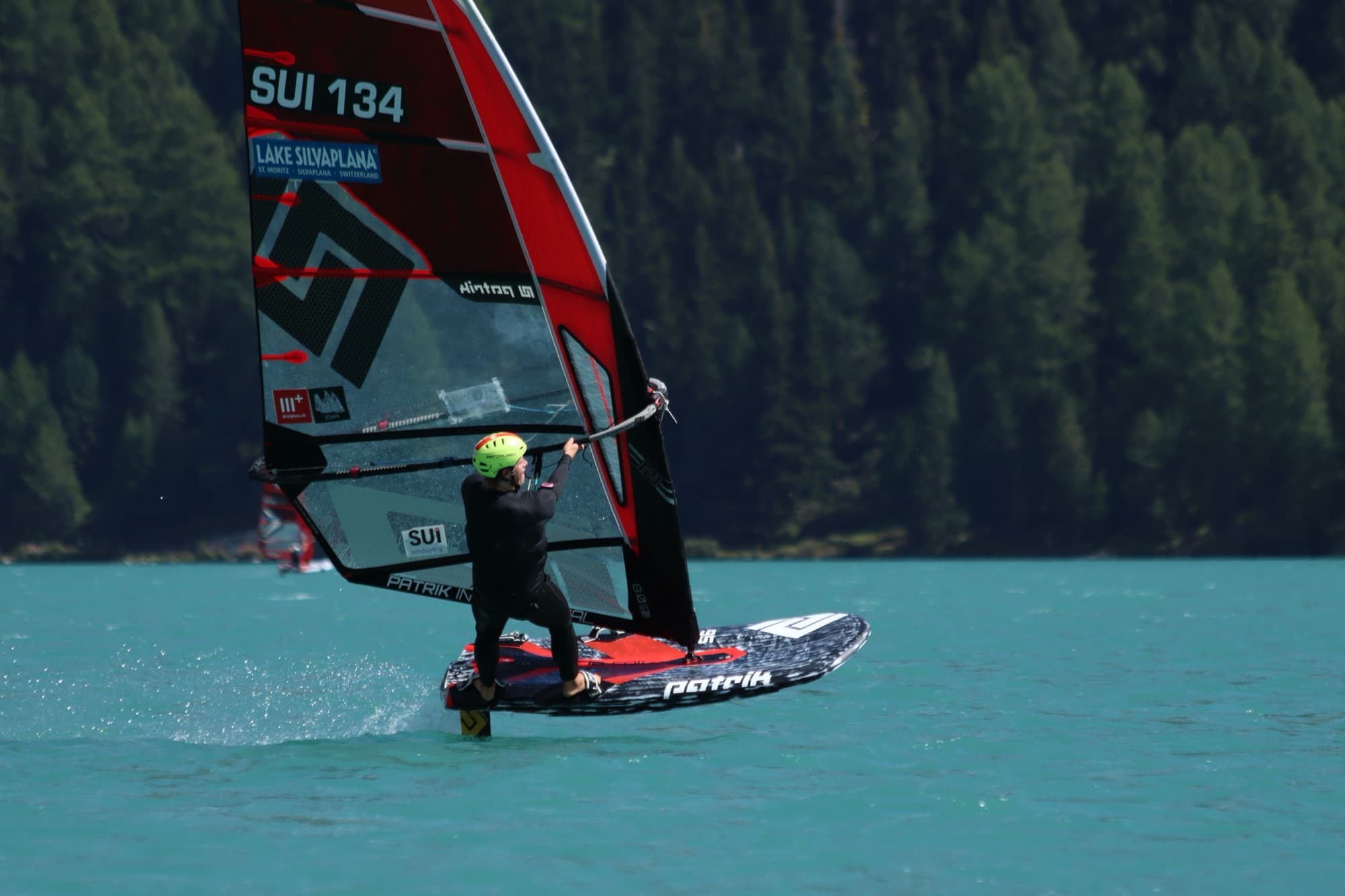 Devin Hauser (SUI 134) auf dem Foil, Board und Rigg ganz im Bild, dunkler Wald dahinter