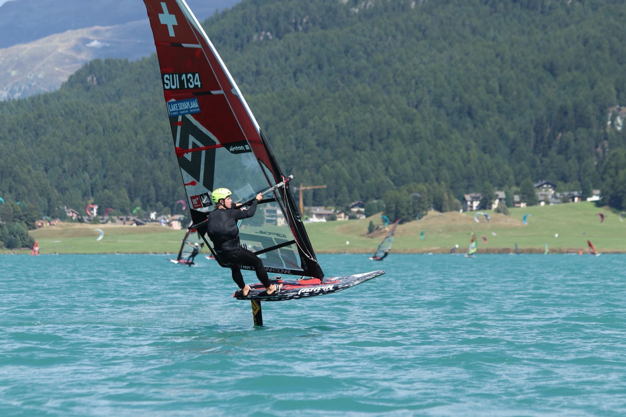 Devin Hauser (SUI 134) auf dem Foil vor Wiesen mit Kites und einem Dorf am Ufer