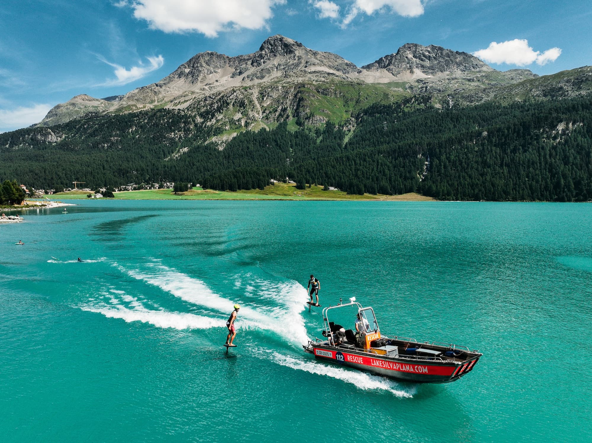 Zwei Fahrer hinter dem roten Rettungsboot auf dem türkisen See, dahinter das Corvatsch-Massiv